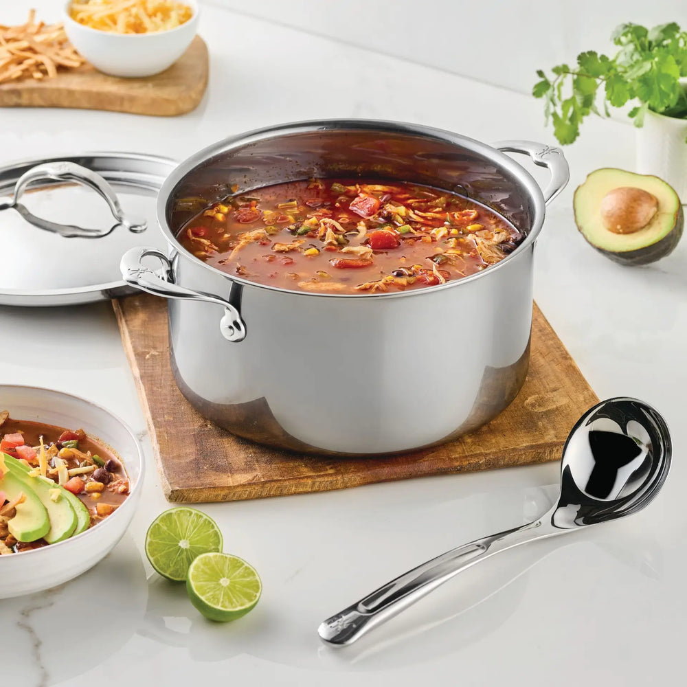 A stainless steel pot of soup with beans, vegetables, and shredded cheese sits on a wooden board. Next to a bowl of soup, the Hestan Provisions 2.4-oz Stainless Steel Serving Ladle rests nearby, while lime halves, cilantro, and a halved avocado are arranged on a white countertop.