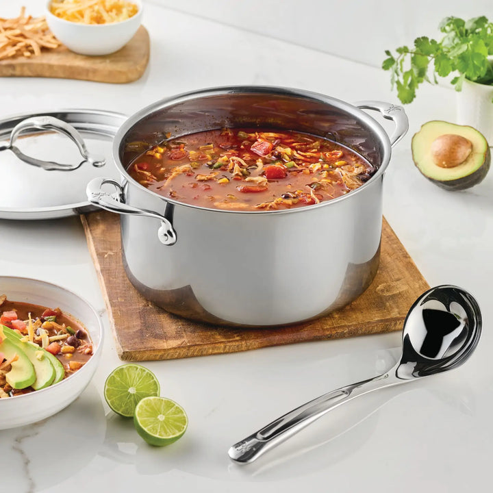 A stainless steel pot of soup with beans, vegetables, and shredded cheese sits on a wooden board. Next to a bowl of soup, the Hestan Provisions 2.4-oz Stainless Steel Serving Ladle rests nearby, while lime halves, cilantro, and a halved avocado are arranged on a white countertop.