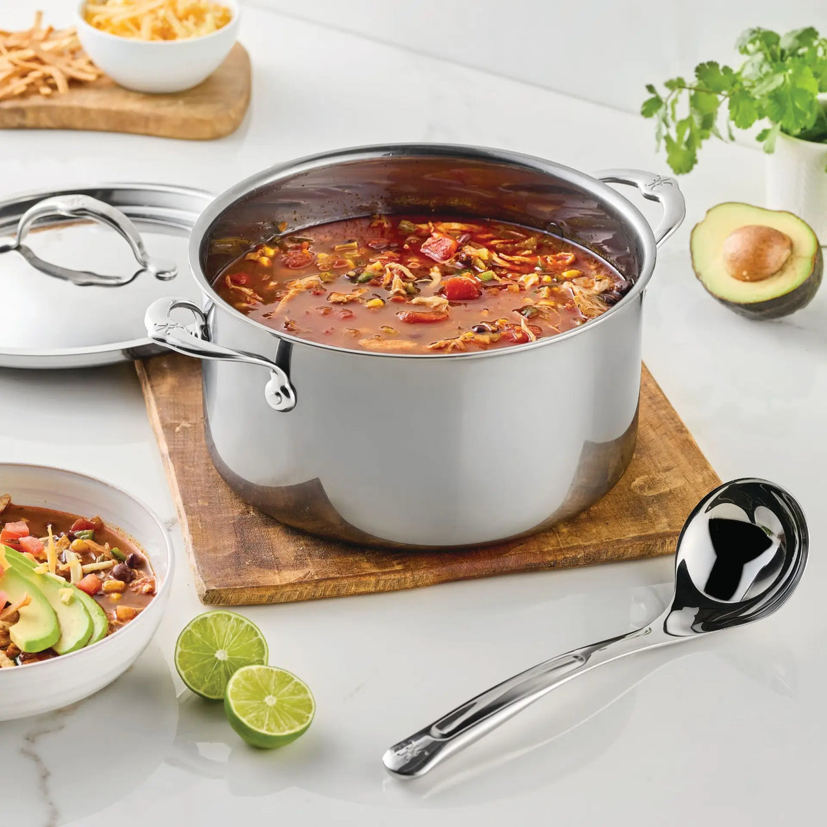 A stainless steel pot of soup with beans, vegetables, and shredded cheese sits on a wooden board. Next to a bowl of soup, the Hestan Provisions 2.4-oz Stainless Steel Serving Ladle rests nearby, while lime halves, cilantro, and a halved avocado are arranged on a white countertop.