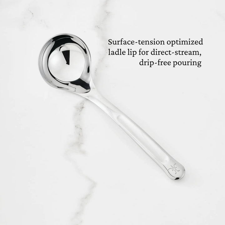 A 2.4-oz Stainless Steel Serving Ladle from the Hestan Provisions collection, featuring a rounded bowl and an engraved handle, rests on a white marble surface. Text on the image highlights its surface-tension optimized lip for direct-stream, drip-free pouring.