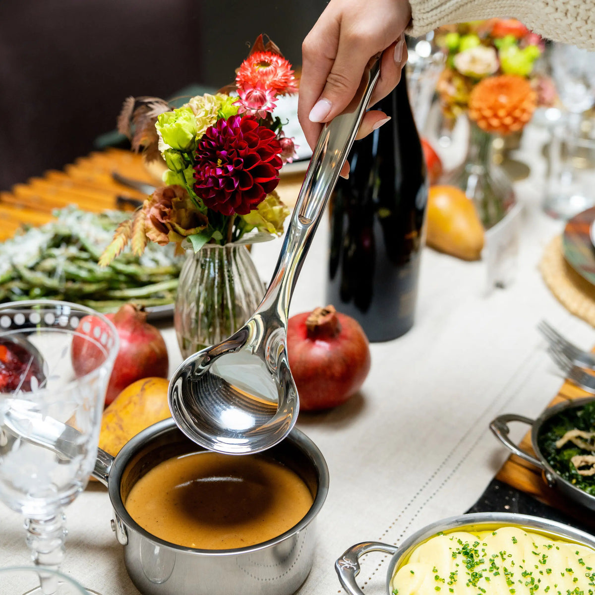 A hand holds a Hestan Provisions® 2.4-oz Stainless Steel Serving Ladle above a small pot of sauce on a set dining table.