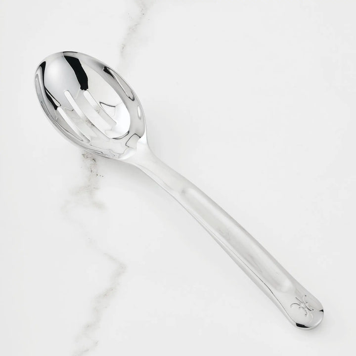 The 10.5-inch Stainless Steel Slotted Serving Spoon from Hestan Provisions rests on a white marble surface. An essential addition to any chef’s toolkit, this spoon features three slots in the bowl and a sleek handle with a subtle engraved detail at the tip.