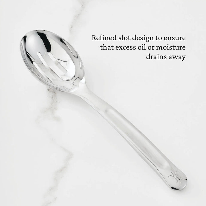 A Hestan Provisions 10.5-inch Stainless Steel Slotted Serving Spoon rests on a white marble surface. Text on the image reads: Refined slot design ensures excess oil or moisture drains away, making it an essential addition to any chef’s tool collection.