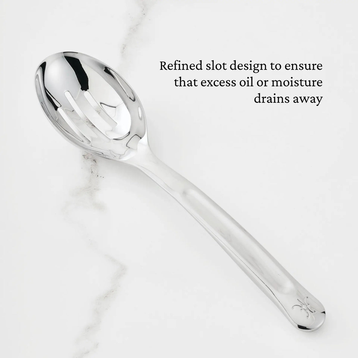 A Hestan Provisions 10.5-inch Stainless Steel Slotted Serving Spoon rests on a white marble surface. Text on the image reads: Refined slot design ensures excess oil or moisture drains away, making it an essential addition to any chef’s tool collection.