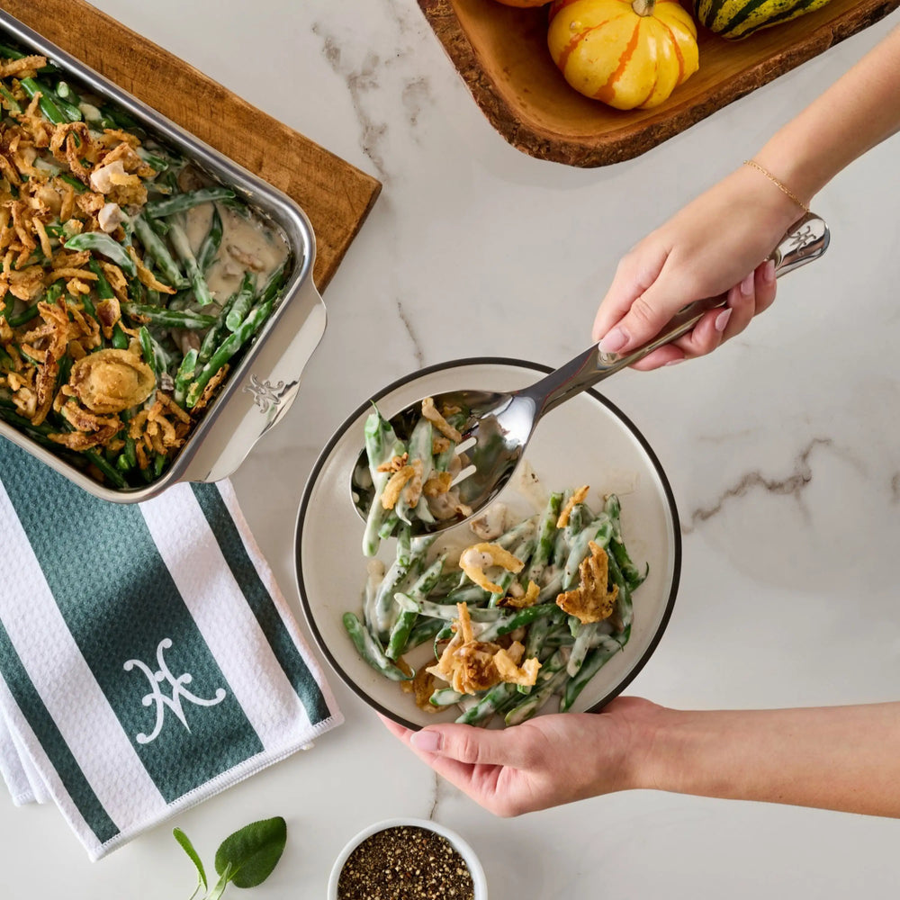 A person uses a 10.5-inch Stainless Steel Slotted Serving Spoon from Hestan Provisions to serve green bean casserole from a baking dish onto a plate.