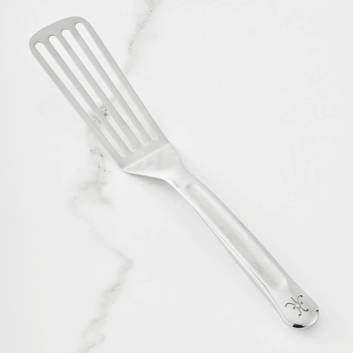 The 11.5-inch Stainless Steel Chef’s Fish Turner from Hestan Provisions, part of their Chef’s Tools collection, features a decorative pattern on the handle and is displayed on a white marble surface.