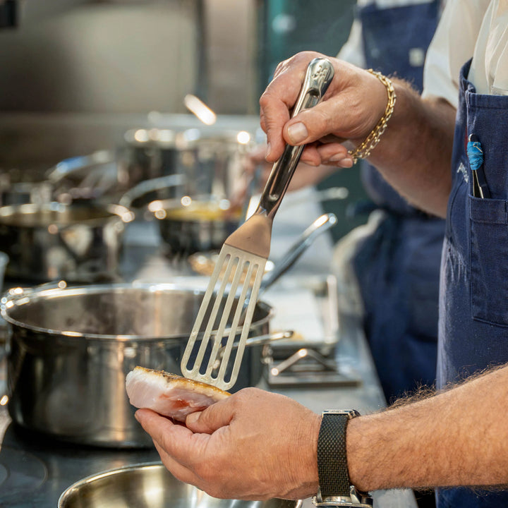 A person in a kitchen holds the Hestan Provisions 11.5-inch Stainless Steel Chef’s Fish Turner and a piece of fish over a pan, with professional kitchen utensils and pots visible in the background. Only the person’s hands and part of their torso are shown.