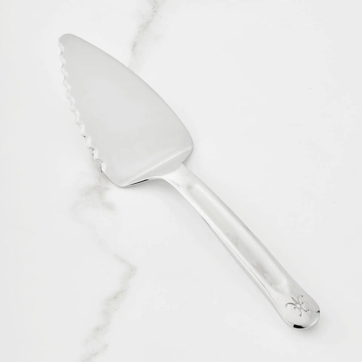 The Hestan Provisions 10.5-inch Stainless Steel Pie & Cake Server, featuring a serrated edge on one side and a decorative emblem on the handle, is displayed on a white marble surface—an elegant addition to any chef’s kitchen tools.