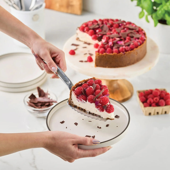 A person serves a slice of cheesecake topped with fresh raspberries and chocolate shavings onto a plate using the Hestan Provisions 10.5-inch Stainless Steel Pie & Cake Server. The remaining cheesecake rests on a cake stand nearby, with raspberries and chocolate scattered on the counter, evoking the elegance of professional chef’s kitchen tools.