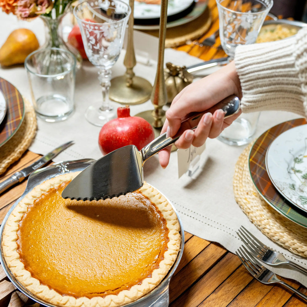 A hand uses a 10.5-inch Stainless Steel Pie & Cake Server from Hestan Provisions® to lift a slice of pumpkin pie from a table set for dinner.