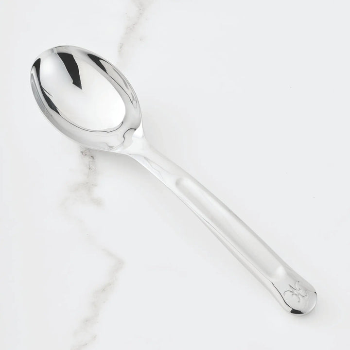 The 10.5-inch Stainless Steel Solid Serving Spoon by Hestan Provisions, an essential chef’s tool, rests on a white surface subtly veined with gray. Its handle is adorned near the end with a delicate decorative emblem.