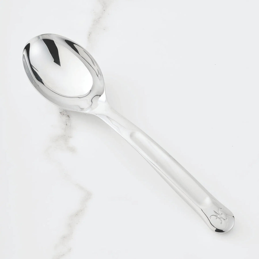 The 10.5-inch Stainless Steel Solid Serving Spoon by Hestan Provisions, an essential chef’s tool, rests on a white surface subtly veined with gray. Its handle is adorned near the end with a delicate decorative emblem.