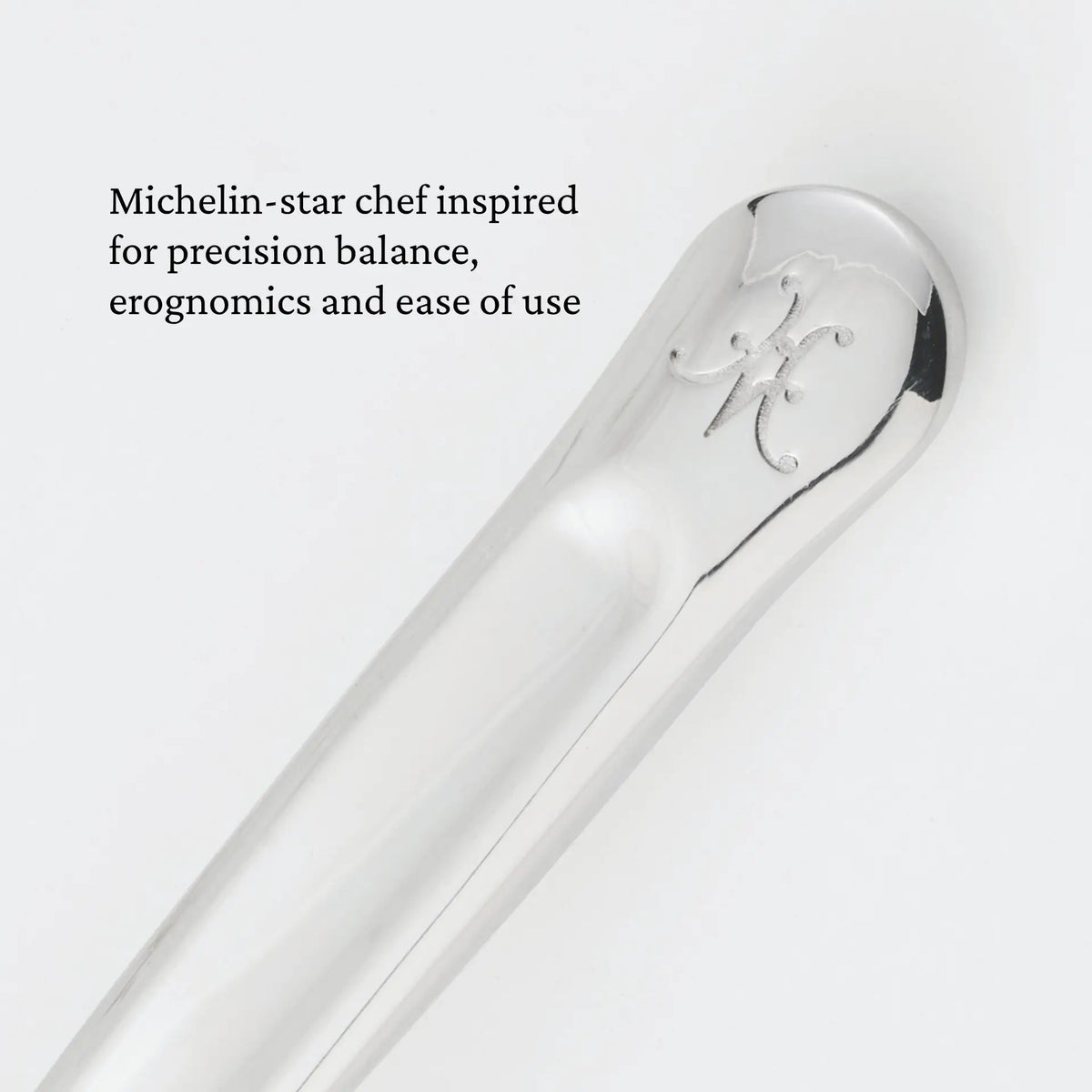 A close-up of a polished metal handle engraved with a stylized M, with text that reads: Inspired by Michelin-star chefs for precision balance, ergonomics, and ease of use—the Hestan Provisions 10.5-inch Stainless Steel Solid Serving Spoon elevates every kitchen with professional-grade performance.