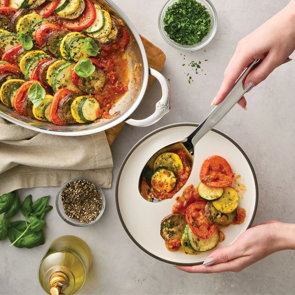 A person serves ratatouille from a white baking dish onto a plate using the Hestan Provisions 10.5-inch Stainless Steel Solid Serving Spoon. The table also holds a bowl of herbs, a pepper grinder, fresh basil, a glass of white wine, and a beige napkin—an inviting scene complete with the essential chef’s tools.