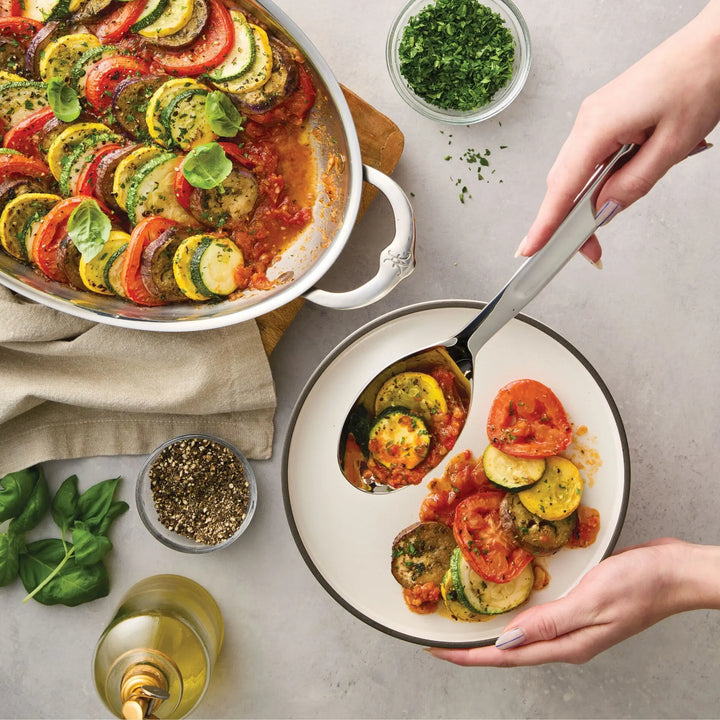 A person serves ratatouille from a white baking dish onto a plate using the Hestan Provisions 10.5-inch Stainless Steel Solid Serving Spoon. The table also holds a bowl of herbs, a pepper grinder, fresh basil, a glass of white wine, and a beige napkin—an inviting scene complete with the essential chef’s tools.