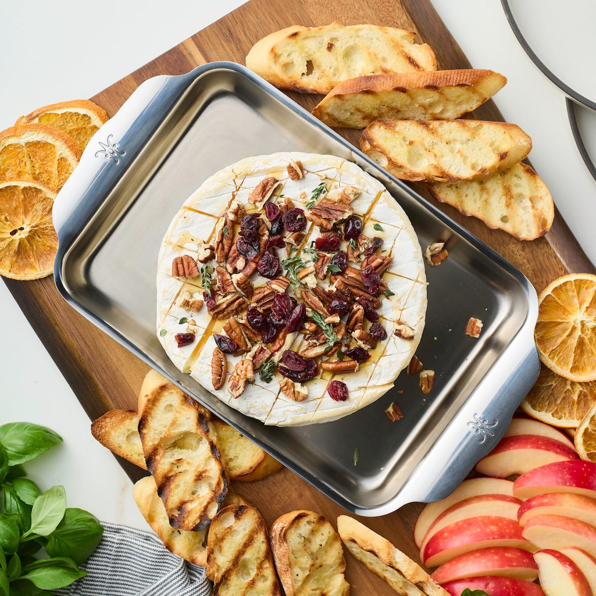 A wheel of brie adorned with pecans, cranberries, and rosemary rests on a Hestan Provisions OvenBond Tri-ply Mini Eighth Sheet Pan with Rack. Its surrounded by sliced baguette, apple slices, dried orange slices, and fresh basil on a wooden board.