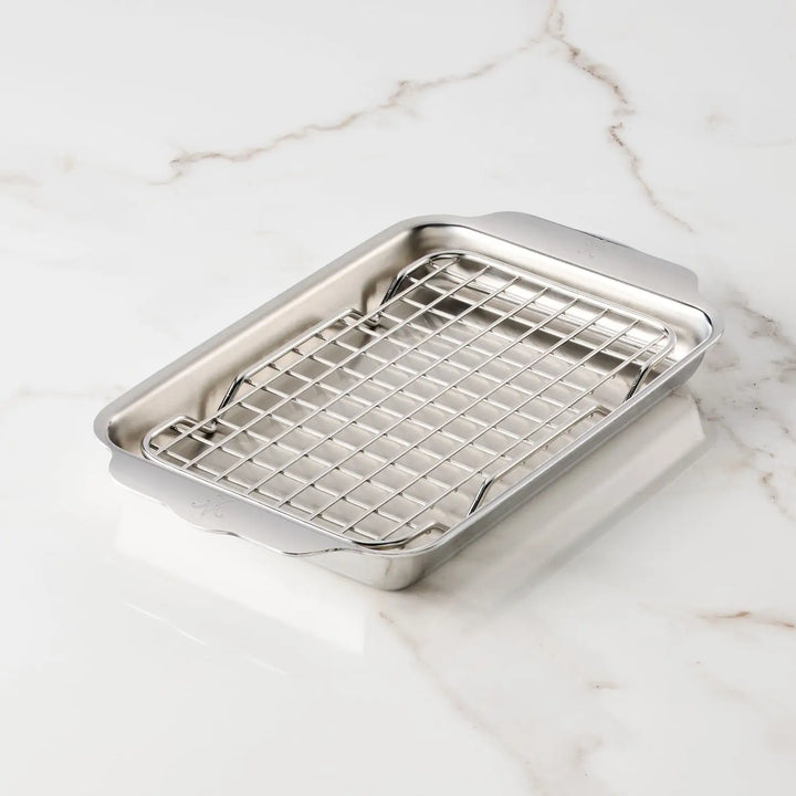 A Hestan Provisions OvenBond Tri-ply Mini Eighth Sheet Pan with Rack elegantly rests on white marble. The stainless steel pan has a built-in roasting rack and features handles on the shorter sides for easy handling.