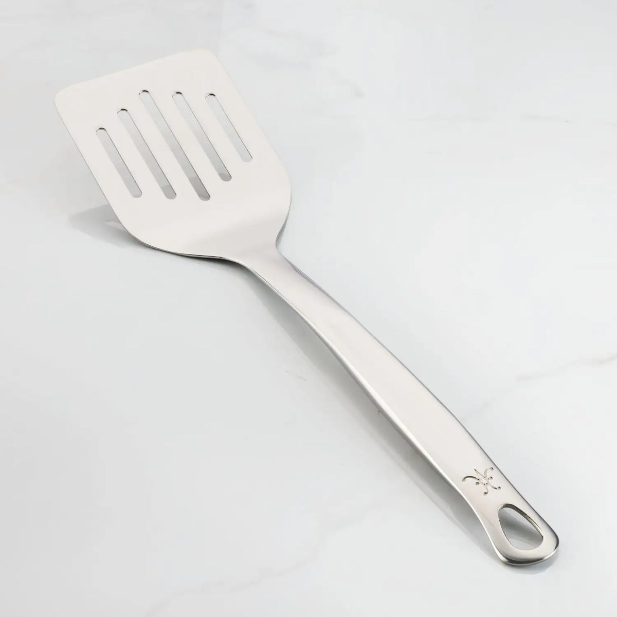 The Hestan Provisions Slotted Turner, 13-inch, crafted from 18/10 stainless steel, features an ergonomic design with a flat rectangular head and long handle with hanging hole, shown resting on a white surface.