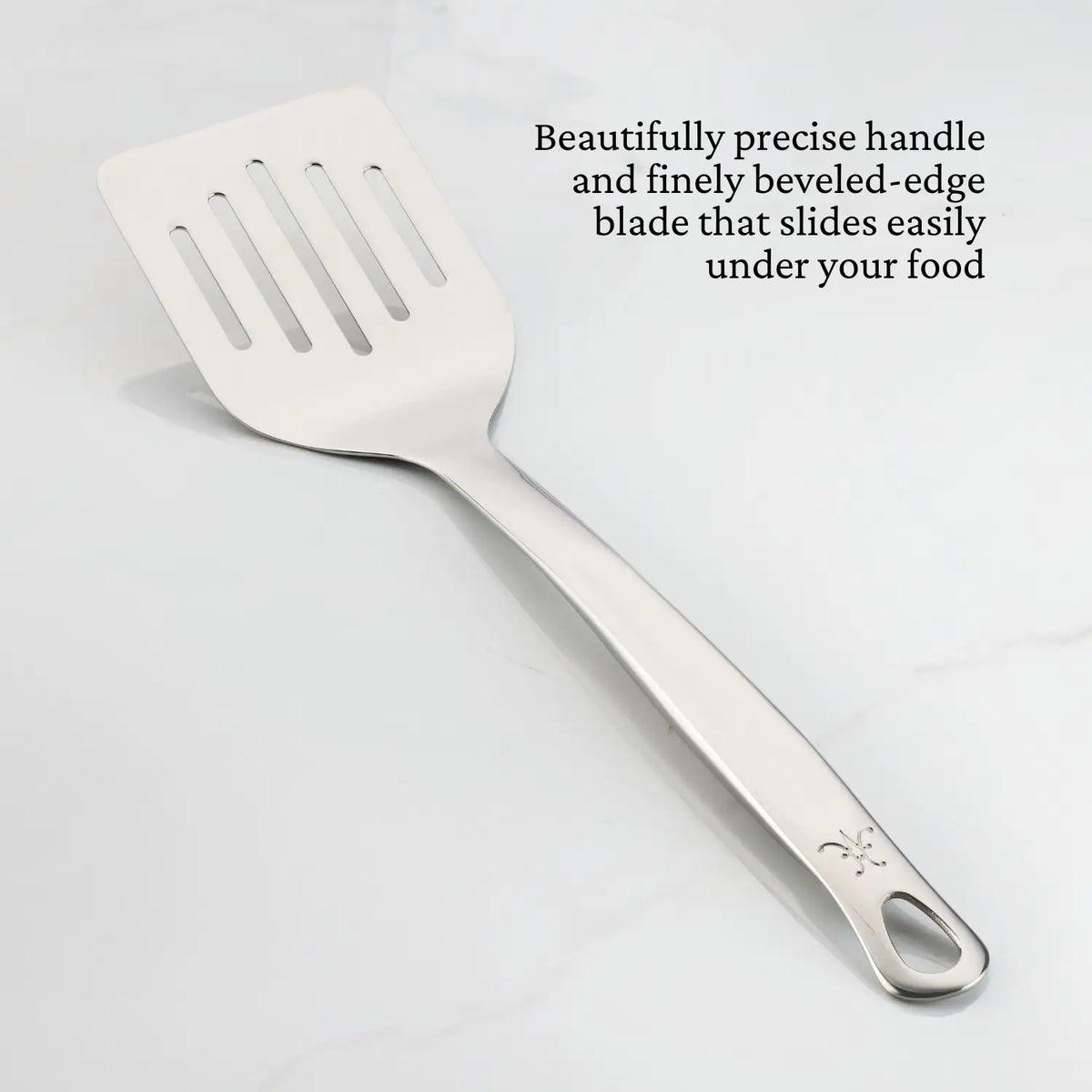 A Hestan Provisions Slotted Turner, 13-inch, with a straight handle rests on a white surface. Text highlights its ergonomic design and finely beveled-edge blade, crafted from 18/10 stainless steel for effortless food handling.