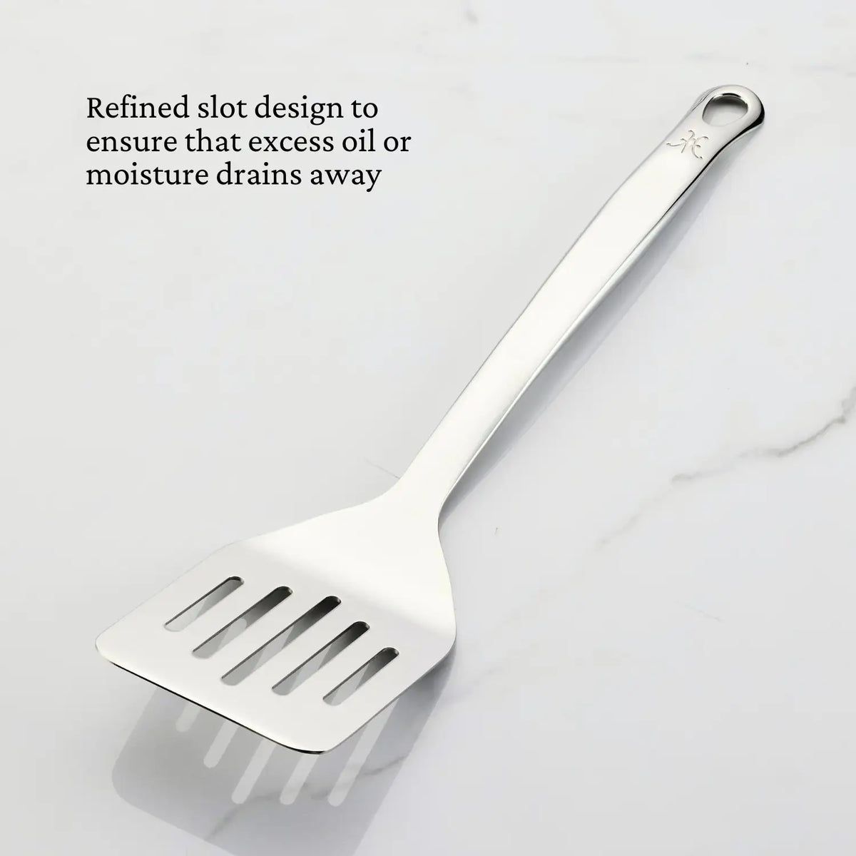 A Hestan Provisions Slotted Turner, 13-inch, made from 18/10 stainless steel with an ergonomic handle and hanging hole, rests on white marble. Text: Refined slots ensure excess oil or moisture drains away.