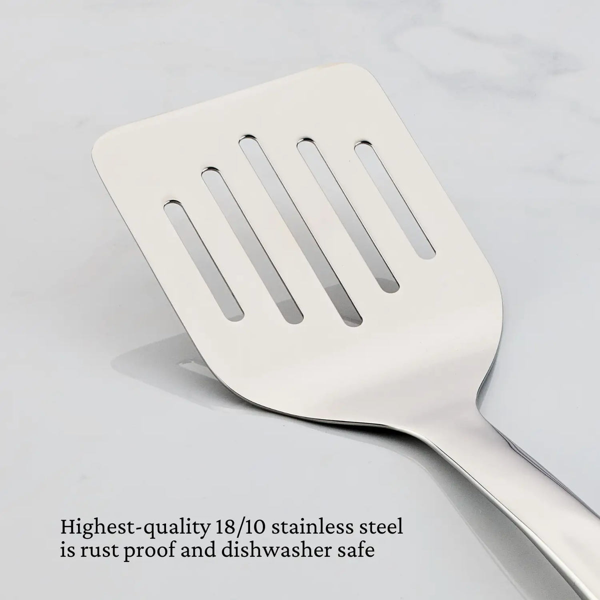 A Hestan Provisions 13-inch Slotted Turner is displayed on a white marble surface. Text below reads, Made from highest-quality 18/10 stainless steel, this ergonomic turner is rust-proof and dishwasher safe.
