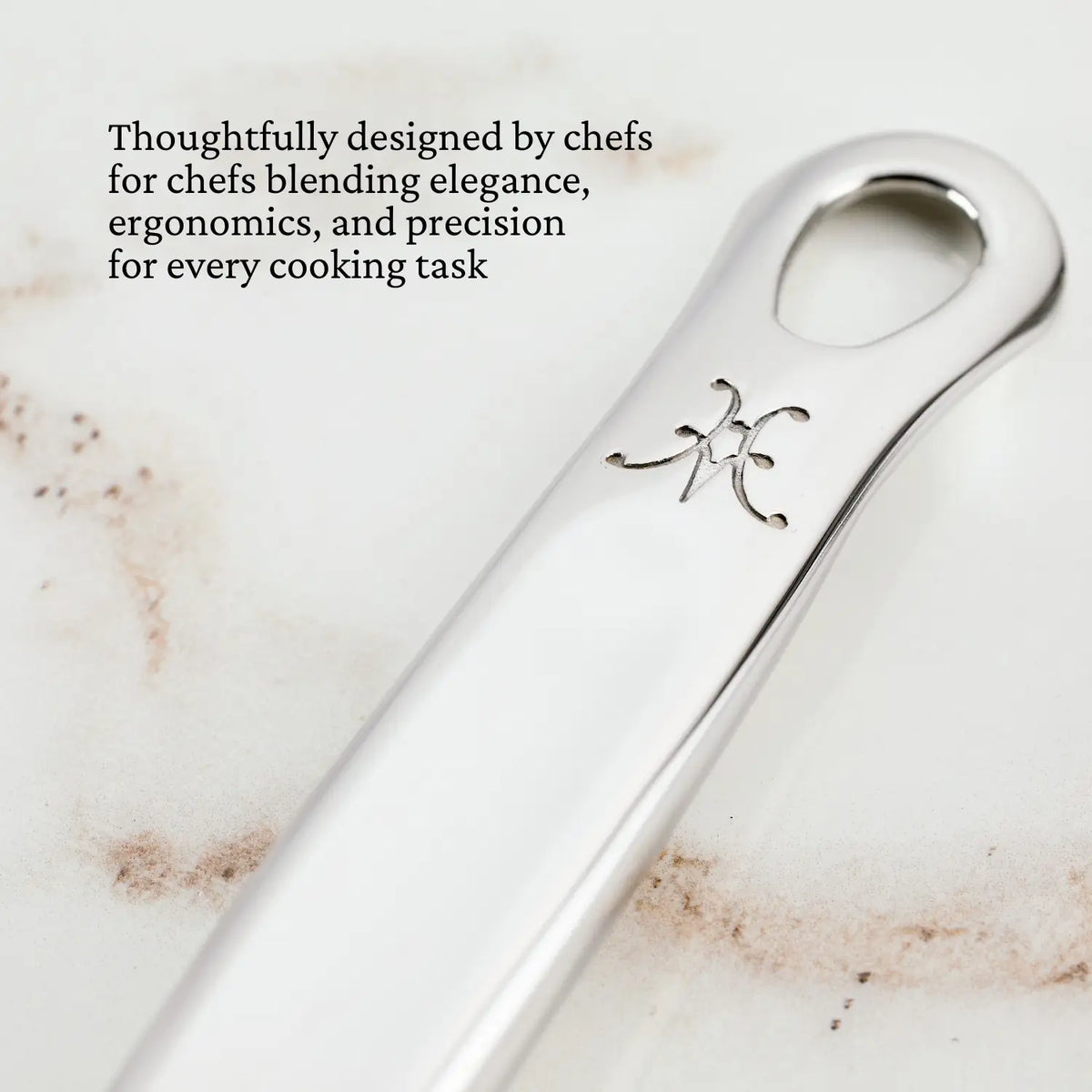 Close-up of a Hestan Provisions Slotted Turner, 13-inch handle in 18/10 stainless steel with an engraved symbol on light marble. Chef-designed for elegance, ergonomic comfort, and precise cooking tasks.