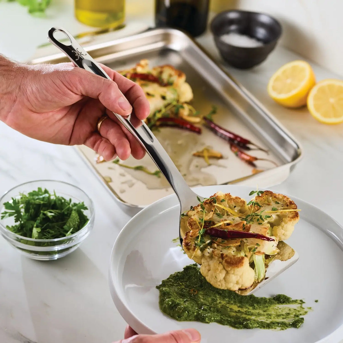A hand uses the Hestan Provisions Slotted Turner, 13-inch to serve a roasted cauliflower steak with herbs and red pepper onto a white plate with green sauce; fresh parsley, lemons, and a baking tray are visible in the background.
