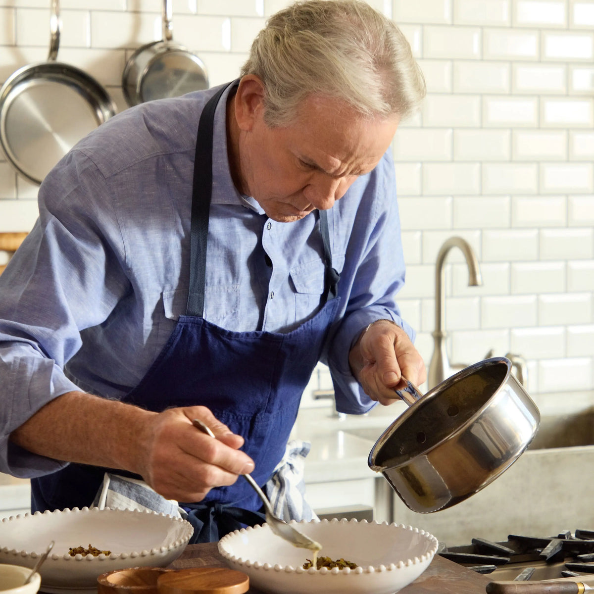 An older man with gray hair and a blue apron focuses on pouring food from his NanoBond Titanium Saucepan into a white bowl on the countertop. Kitchen utensils hang on a tiled wall, emphasizing the cookwares metal utensil-safe feature.