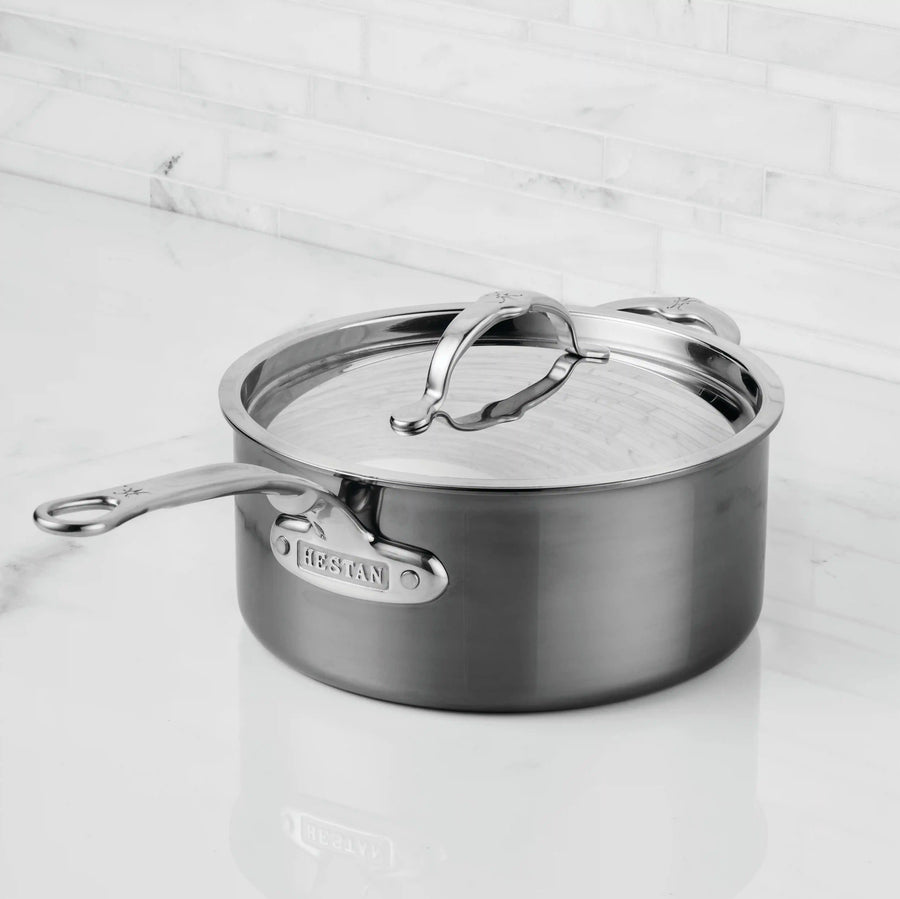 A NanoBond Titanium Saucepan with sturdy handles sits on a white marble countertop, its lid slightly tilted to reveal the interior and showcase its innovative finish for enhanced durability and a sleek look.