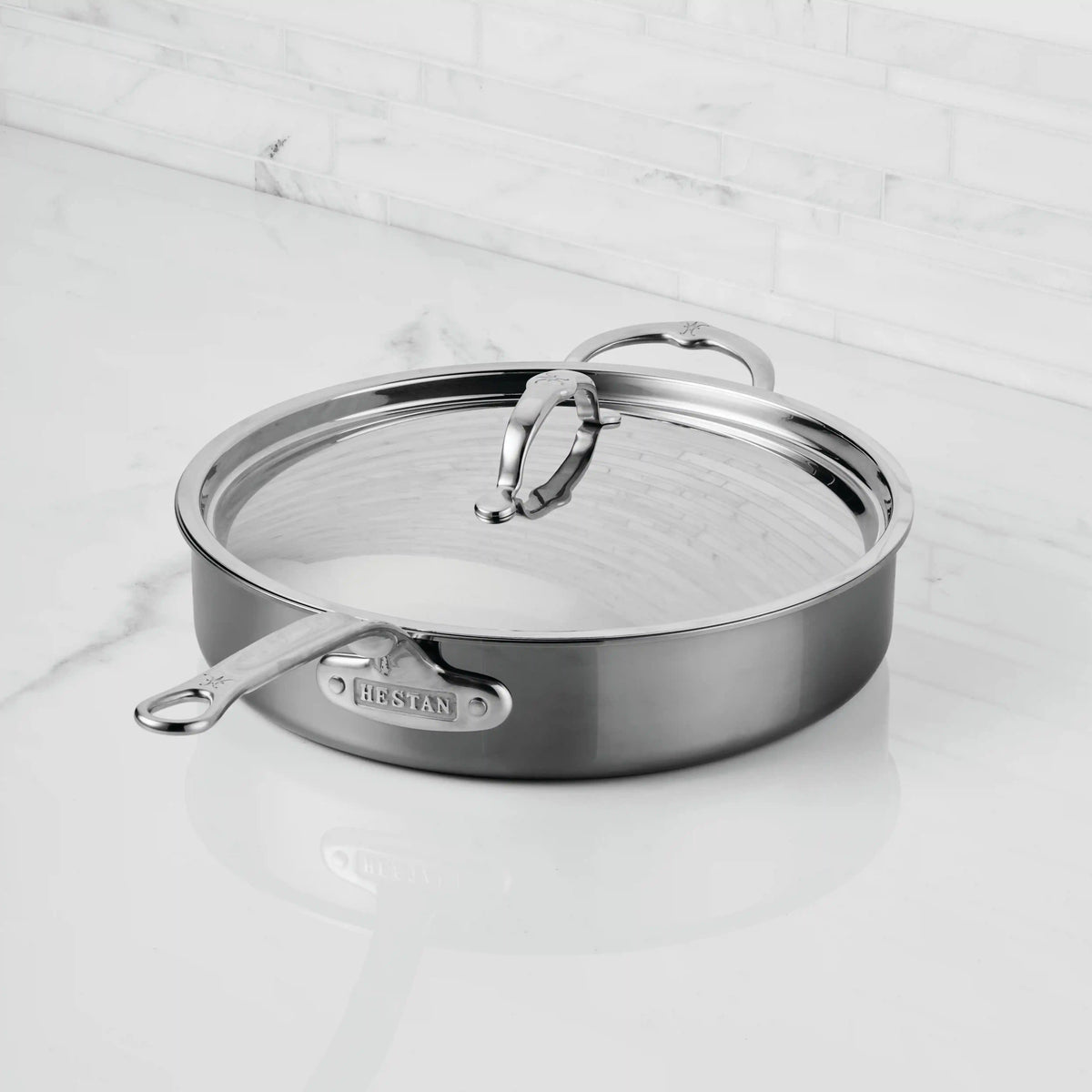 A NanoBond Titanium Sauté Pan with lid sits on a white marble countertop, featuring two handles and a brand label. Its stainless steel surface is strengthened with Molecular Titanium for outstanding durability.