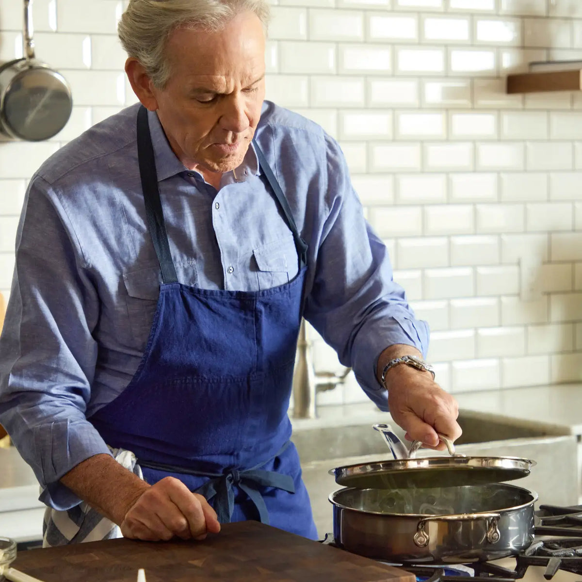 An older man in a blue apron and shirt cooks in a kitchen, stirring food in a NanoBond Titanium Sauté Pan on the stove. The kitchen features white subway tile walls and a metal cookware hanger, reflecting his commitment to quality and style.