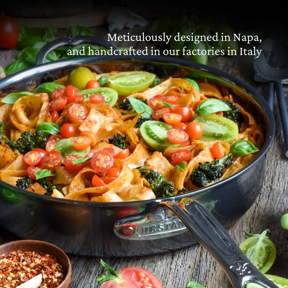 A NanoBond Titanium Sauté Pan holds pasta with tomato sauce, topped with cherry tomatoes, sliced green tomatoes, and basil leaves. In the background are herbs and a bowl of crushed red pepper. Text above notes design in Napa and Italian craftsmanship.