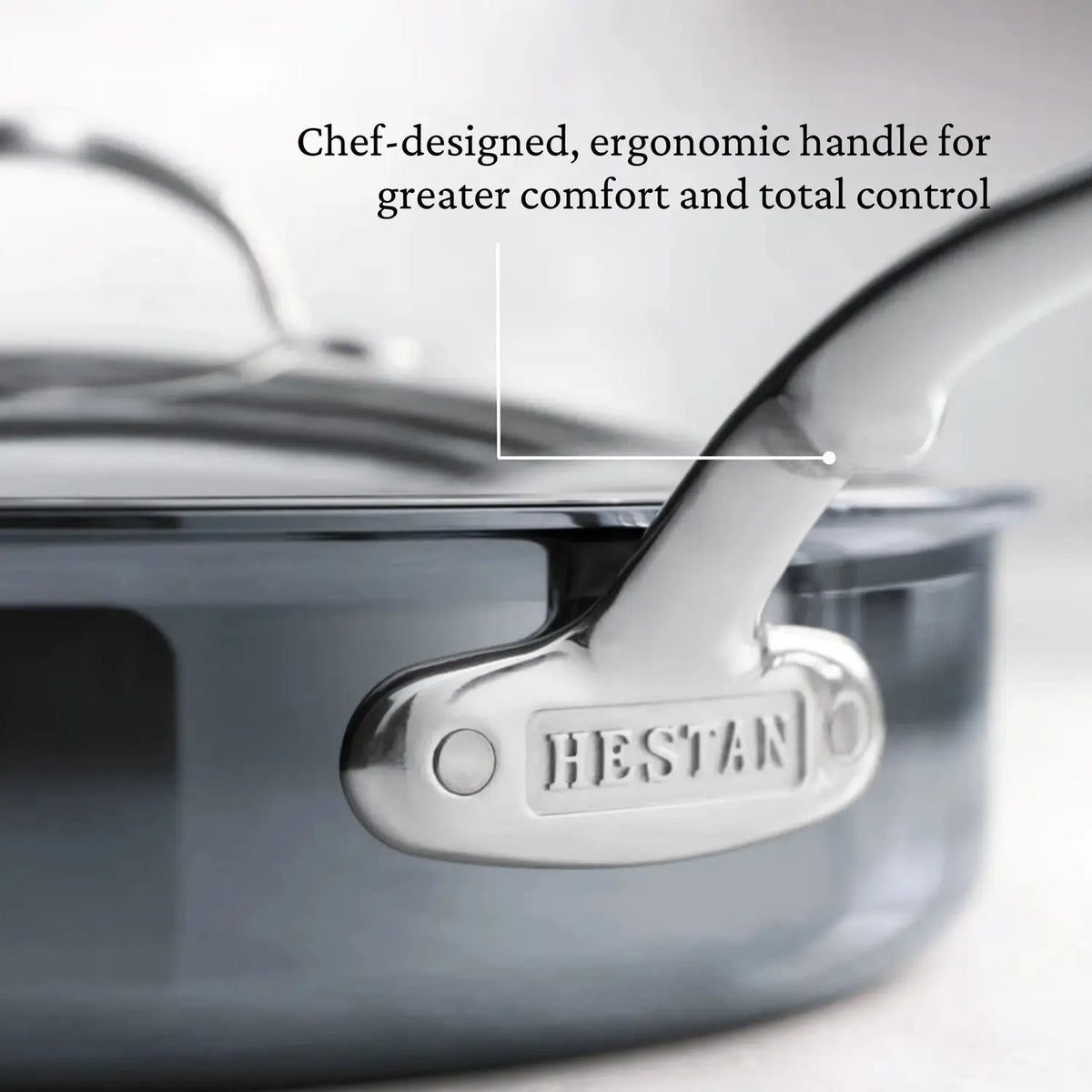 A close-up of a metal saucepan handle embossed with HESTAN highlights the chef-designed, ergonomic design for comfort and control. The NanoBond Titanium Sauté Pan features a polished finish, crafted with advanced Molecular Titanium technology for enhanced durability.