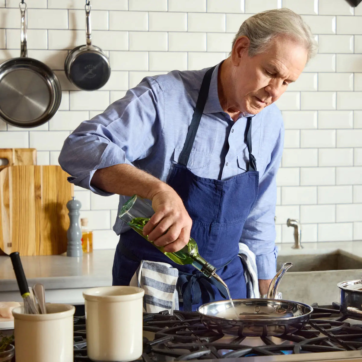 An older man in a blue shirt and apron pours olive oil into a NanoBond Titanium Skillet on a stove in a kitchen with white subway tile, hanging pans, and utensils nearby.