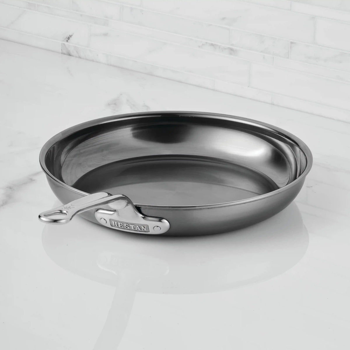 A NanoBond Titanium Skillet with a stainless finish and metal handle sits on a white marble countertop, backed by a white tiled backsplash.
