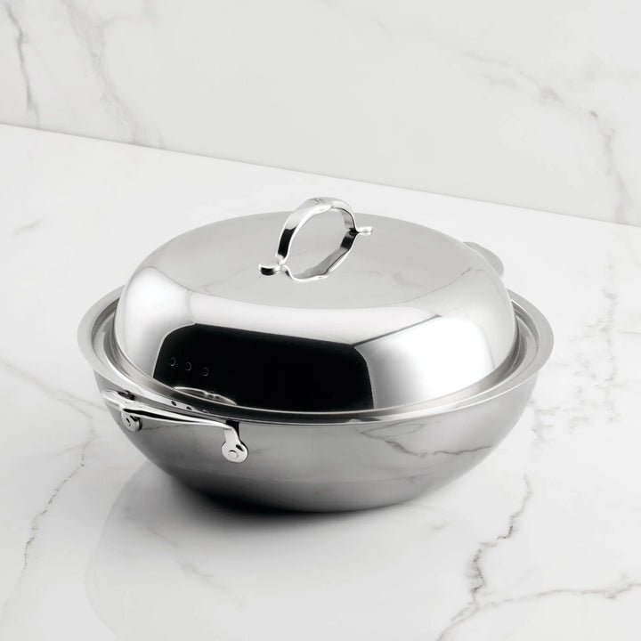 The NanoBond Titanium Wok, 14-Inch, with a lid and side handle, rests on a white marble surface. Made from high heat resistant materials, it features a shiny, reflective finish and a sleek modern design.