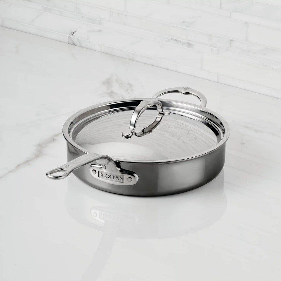 A NanoBond Titanium Sauté Pan with a lid and dual handles rests on a white marble countertop, set against a white tiled backsplash.