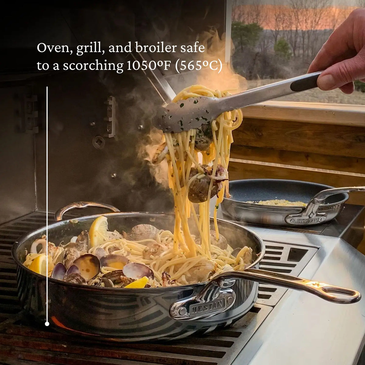 Using tongs, someone lifts pasta with clams and lemon from a steaming NanoBond Titanium Sauté Pan on the grill. Text reads, Safe for oven, grill, and broiler up to 1050°F (565°C). Another NanoBond Titanium Pan is in the background.