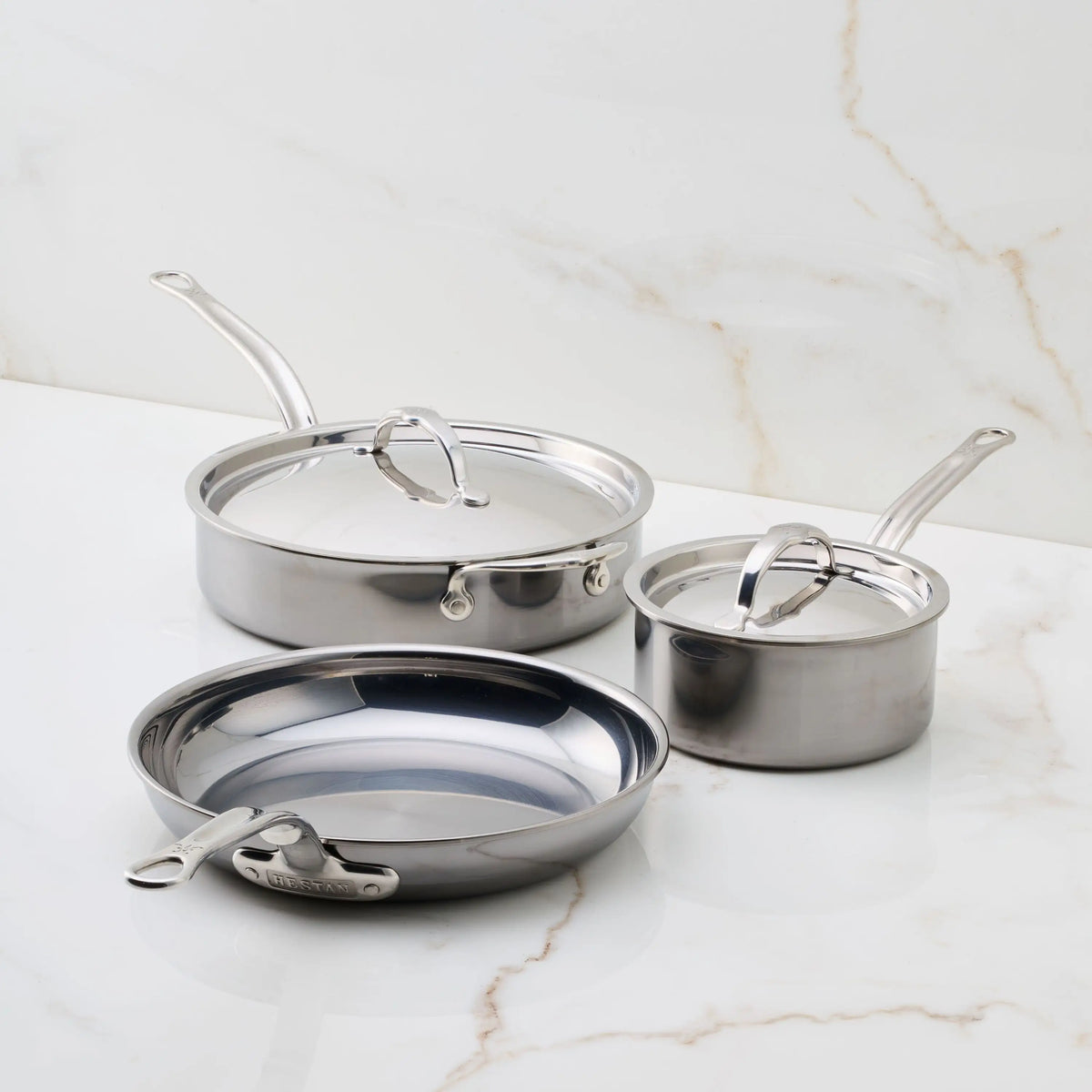 Three pieces from the NanoBond 5-Piece Titanium Essential Cookware Set—a frying pan, covered saucepan, and covered sauté pan—are arranged on a white marble surface.