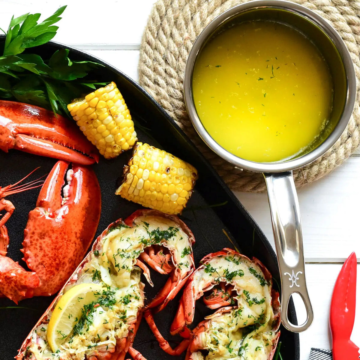 A cooked lobster with two corn cob slices rests on a black tray, accompanied by a NanoBond Titanium Butter Warmer, .75 quarts, filled with herb-garnished melted butter. Parsley sprigs adorn the dish, and a red claw cracker lies nearby on the white surface.