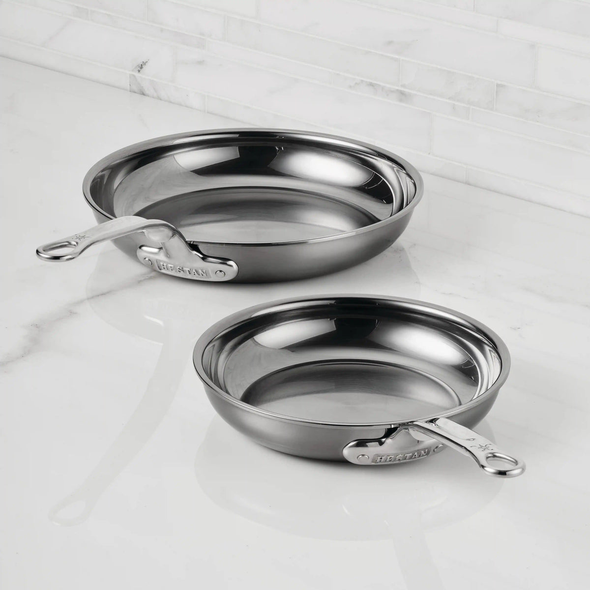 Two NanoBond Titanium Skillets with handles, including a premium titanium skillet, rest on a white marble countertop in a kitchen with a white tiled backsplash.
