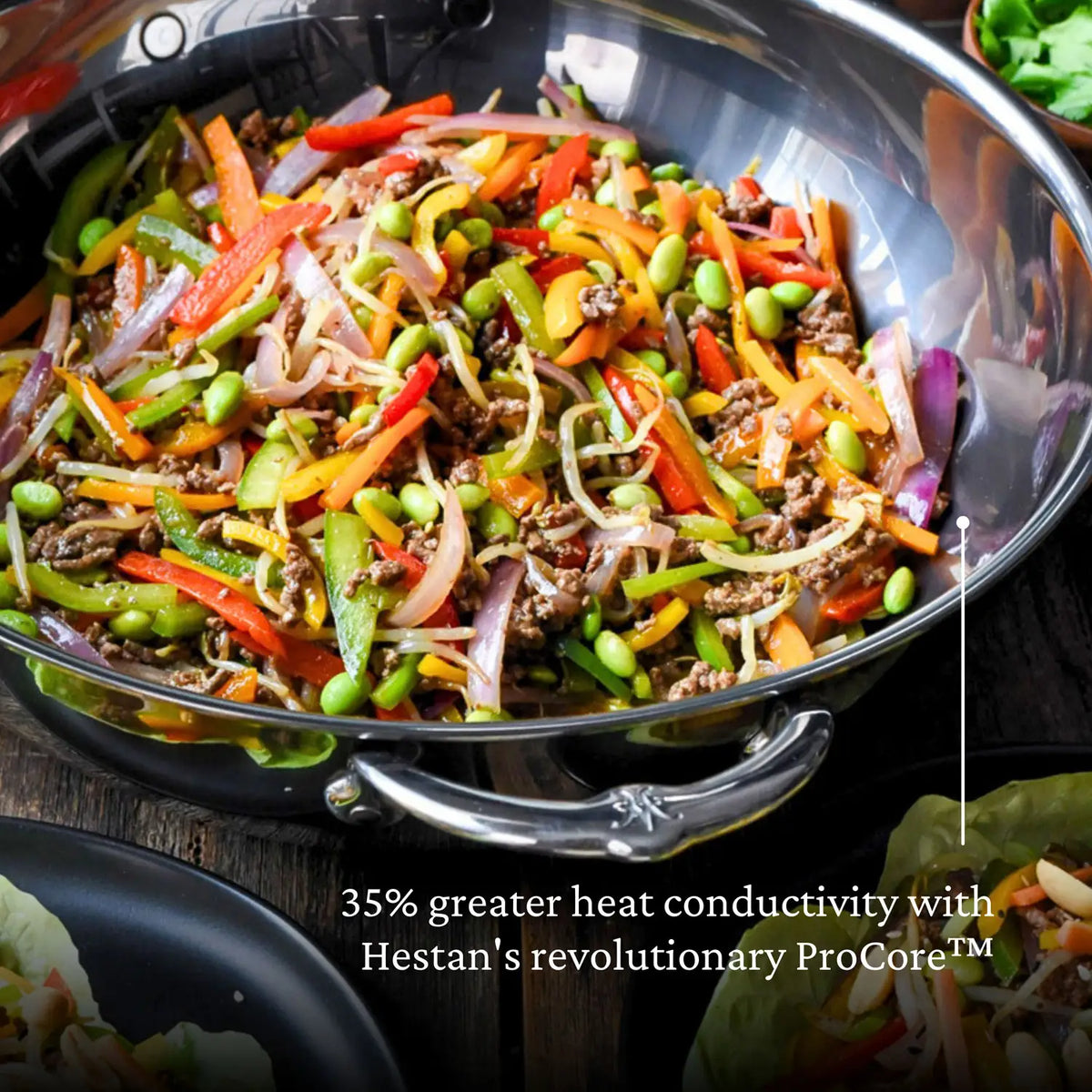 A stir-fry with bell peppers, onions, carrots, edamame, and beef shines in a silver 14-inch Titanium Chefs Pan by NanoBond. The image showcases its innovative ProCore™, providing 35% greater heat conductivity. A lettuce leaf peeks from the side.