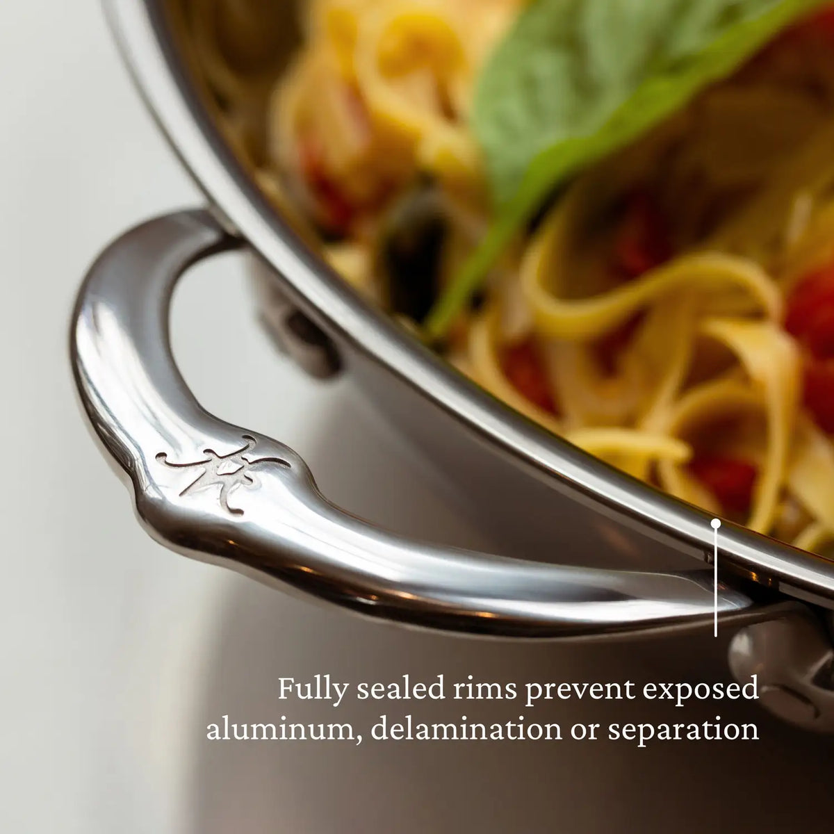 Close-up of a stainless steel pot handle with logo, showing pasta with tomatoes and basil. Text overlay: NanoBonds 14-Inch Titanium Chefs Pan features Molecular Titanium tech for fully sealed rims, preventing exposed aluminum, delamination, or separation.