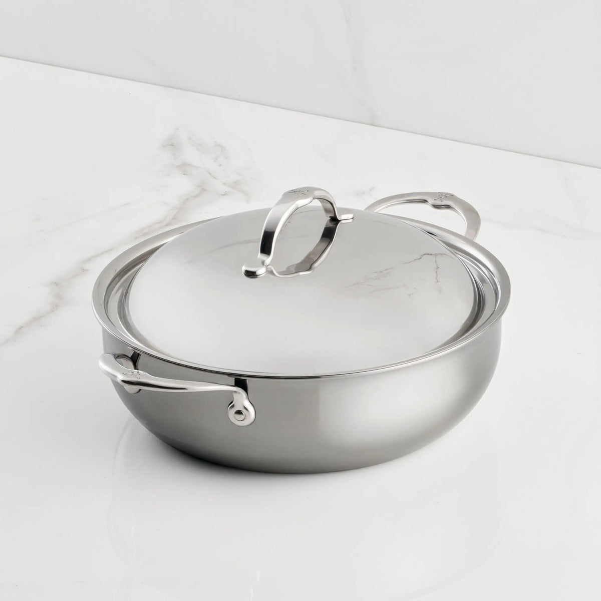 A NanoBond Titanium Dutch Oven, 5-Quart, with two handles and a loop-handled lid rests on a white marble countertop. Its highly reflective stainless steel surface enhances the sleek look.