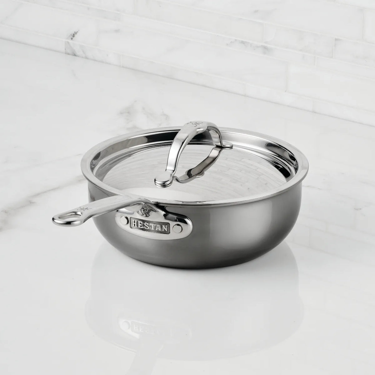 The NanoBond Titanium Stainless Steel Saucier Pan, 2-Quart, with a fitted stainless steel lid, gleams on a white marble surface. It features a side handle and branded lid handle for easy lifting.