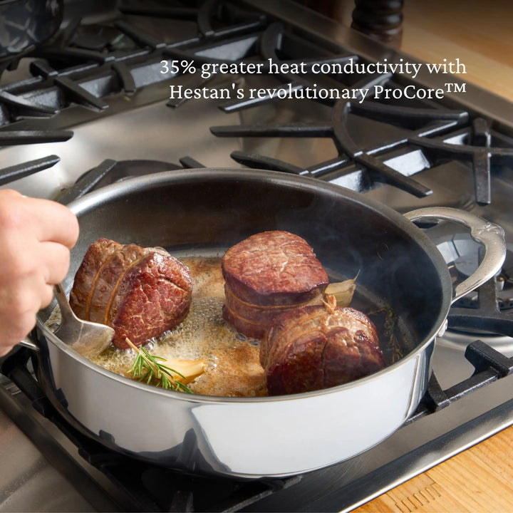 A hand uses tongs to sear three pieces of beef in a NanoBond Titanium Stockpot on a gas stove. Text above the pot reads: 35% greater heat conductivity with Hestan’s revolutionary Molecular Titanium Cookware.