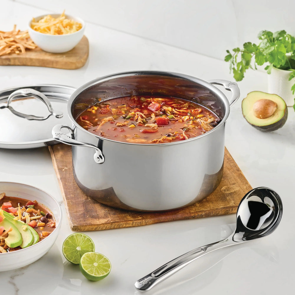 A NanoBond Titanium Stockpot filled with soup sits on a wooden board, surrounded by a ladle, lime halves, a bowl of soup, sliced avocado, shredded cheese, and cilantro on a white countertop.