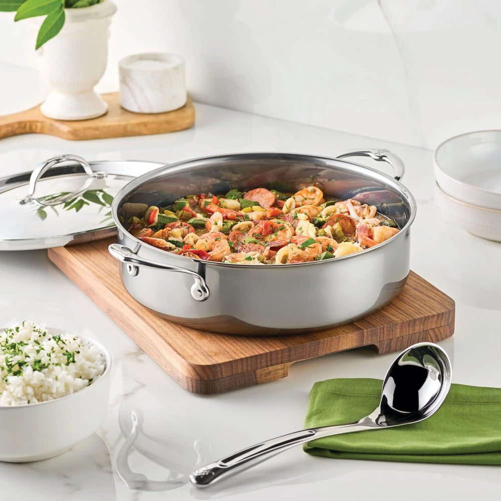 A NanoBond Titanium Rondeau filled with mixed stir-fry sits on a wooden cutting board, next to a bowl of rice, serving spoon on a green napkin, and a marble mortar and pestle atop a white kitchen counter.