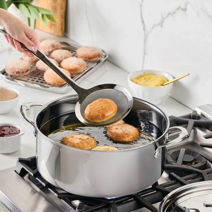 A hand uses a slotted spoon to lift a fried donut from a NanoBond Titanium Rondeau on the stove. More donuts fry in the pot, while finished ones cool nearby. Bowls of condiments rest on the counter.