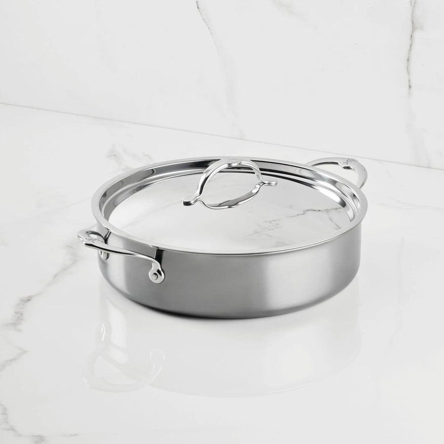 A NanoBond Titanium Rondeau with two handles and a matching lid rests on white marble with grey veins, featuring a shiny, reflective finish and a sleek, modern look.